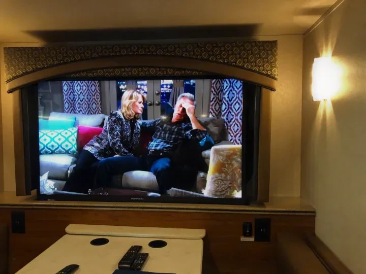 Wall-mounted TV in the living area playing a show, with reading lamp lit alongside