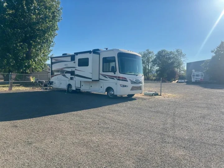 Driver-side exterior view of the motorhome parked behind a fence with slide-outs visible