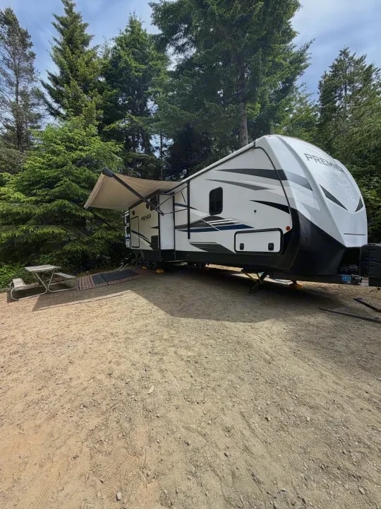 Trailer with awning extended over a picnic table and rug at a wooded campsite