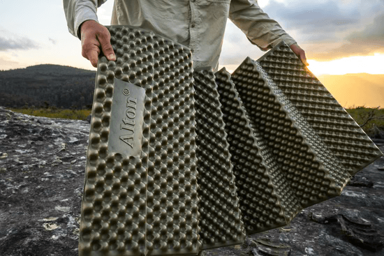 Camper unfolding a green closed-cell foam sleeping pad on a rock at sunset