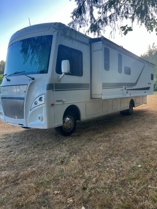 Front three-quarter view of the Winnebago Intent motorhome with awning extended under trees