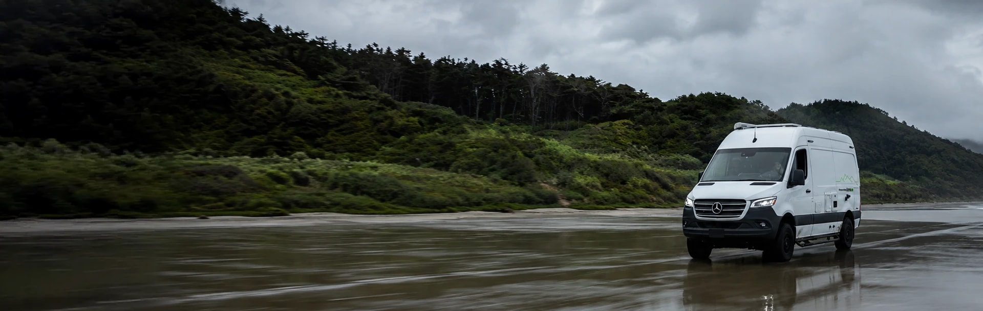A camper van driving along a wet sand beach beneath forested hills