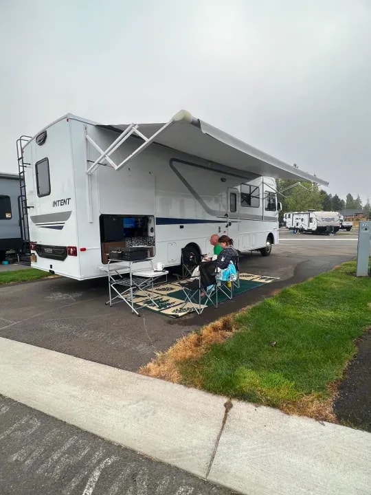 Motorhome parked at a campsite with awning out and two people seated at a folding table